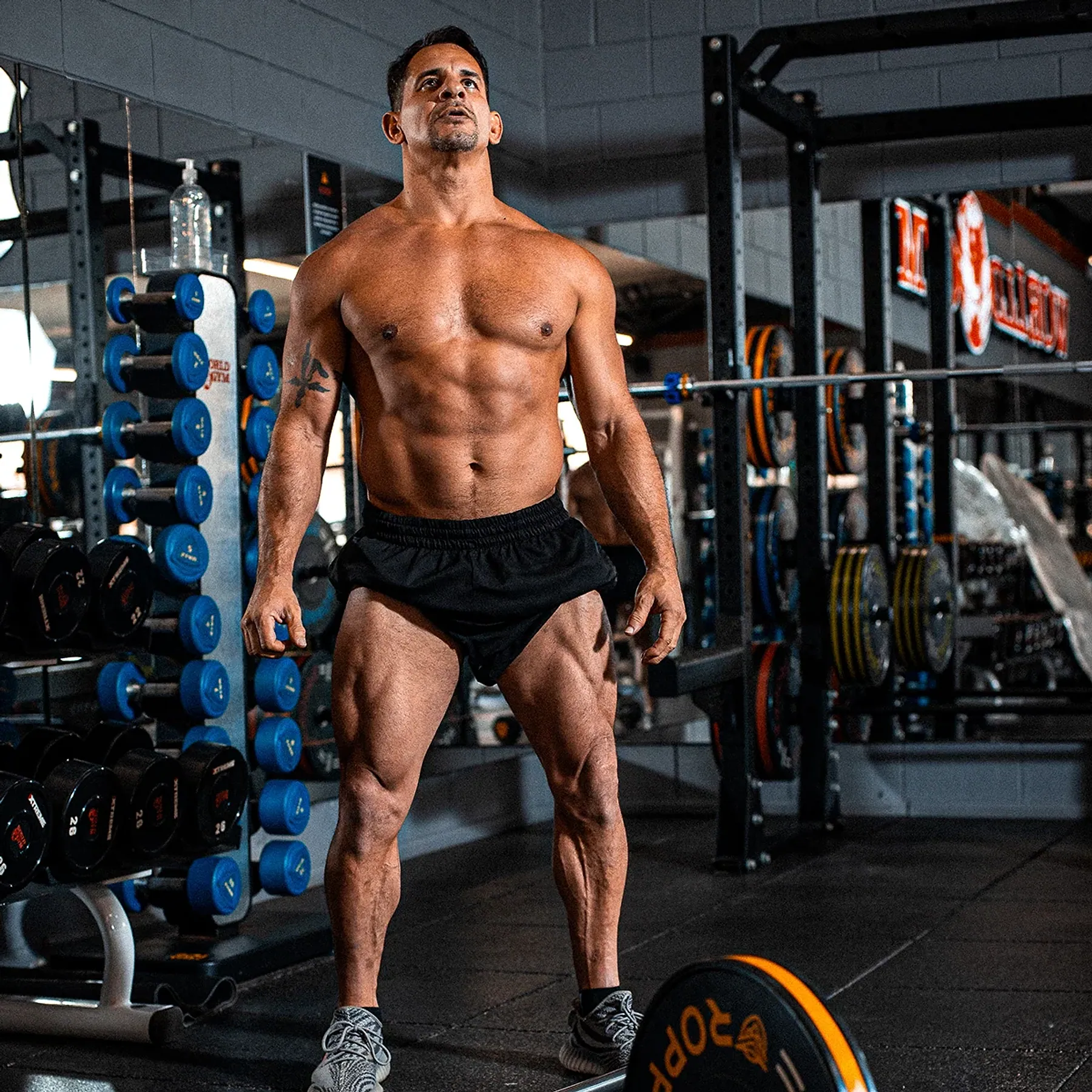 Chico em pé, de corpo inteiro, em uma academia com racks e halteres ao fundo.