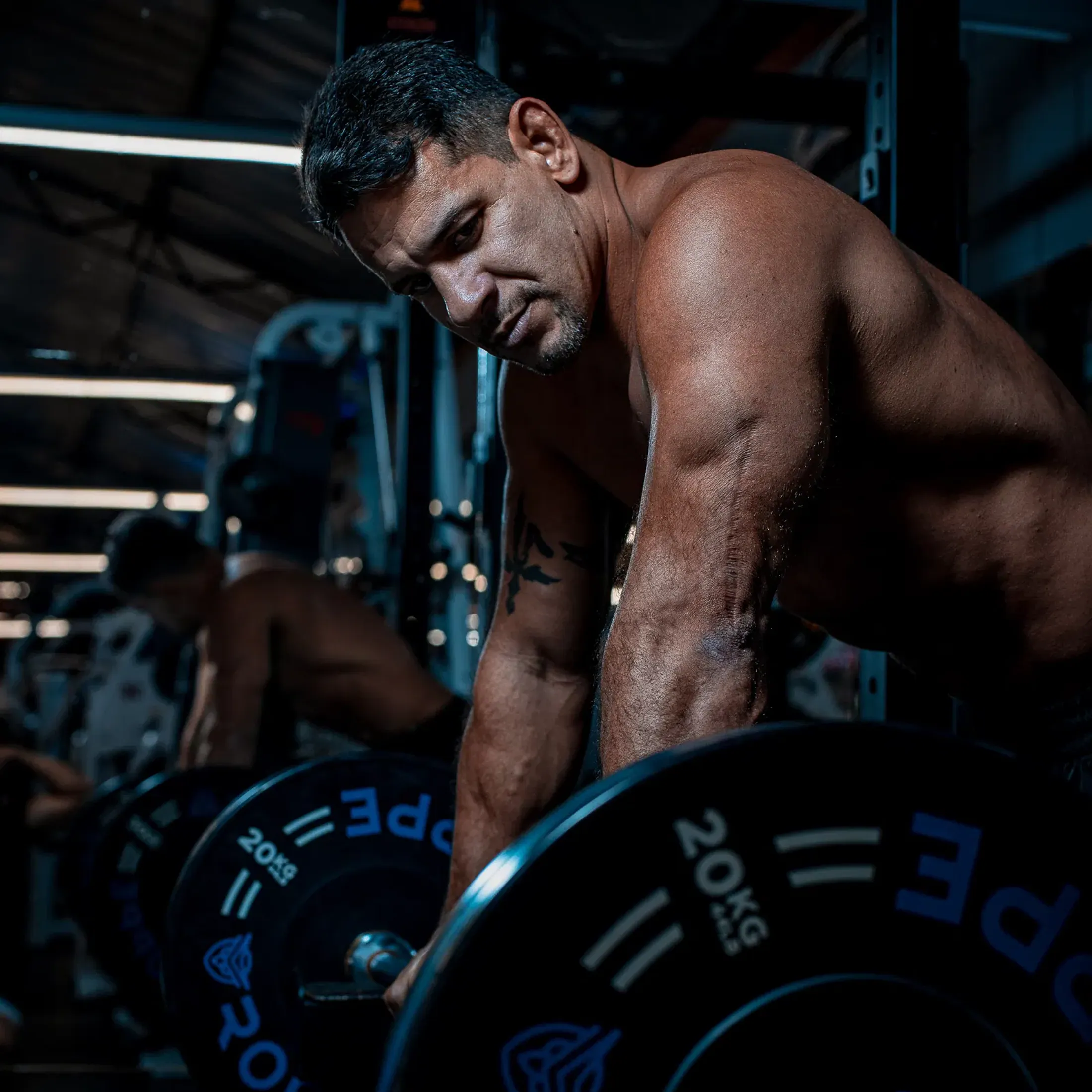 Chico debruçado sobre uma barra carregada, de cabeça baixa, em uma academia com luz fria.